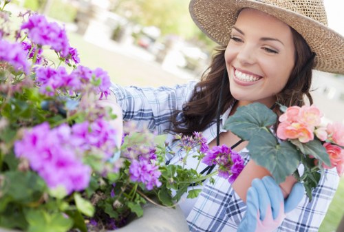 A woman in a straw hat and light blue gloves is engaged in gardening within a well-maintained outdoor space, likely a front or backyard garden in East Ham. She is smiling as she tends to a potted flower arrangement that features vibrant purple and soft pink blossoms, with lush green foliage. In the background, a neatly mowed lawn with dense, green grass stretches across the garden area, bordered by a paved patio or pathway made of textured concrete or stone slabs. The garden includes a variety of plants and flowering pots, with some shrubs and small trees visible further back, contributing to a layered landscape design typical of residential outdoor spaces in East London. The natural lighting indicates a bright, clear day, enhancing the colours of the flowers and greenery. The scene exemplifies professional gardening services focused on floral care and outdoor maintenance, subtly aligned with local gardening standards and landscape aesthetics, with the context rooted in an East Ham postcode area. This photograph highlights thorough garden care, including planting and nurturing flowering plants in a tidy outdoor environment.