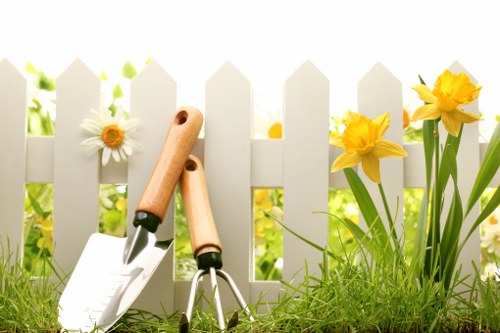 A close-up image of gardening tools, including a pair of hand trowels with wooden handles and a garden fork with a metal head, resting against a white picket fence. The foreground features a lush green lawn with well-maintained grass and a few small yellow and white flowers growing near the fence. Behind the fence, there are taller yellow daffodils and white daisies, adding vibrant colour to the scene. The background shows a blurred outdoor garden setting with greenery suggesting a sunny day, suitable for outdoor gardening and lawn care activities commonly undertaken by gardening services such as Hedge Trimming East Ham. This image captures the elements typically involved in garden maintenance, from planting to trimming, within a tidy, well-kept outdoor space that may be representative of a suburban garden in East Ham, London.