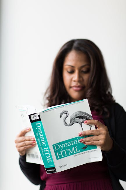 A woman with dark brown hair, dressed in a black blazer and a dark pink top, is standing indoors against a plain white background. She is holding an open book titled 'Dynamic HTML', which features a d