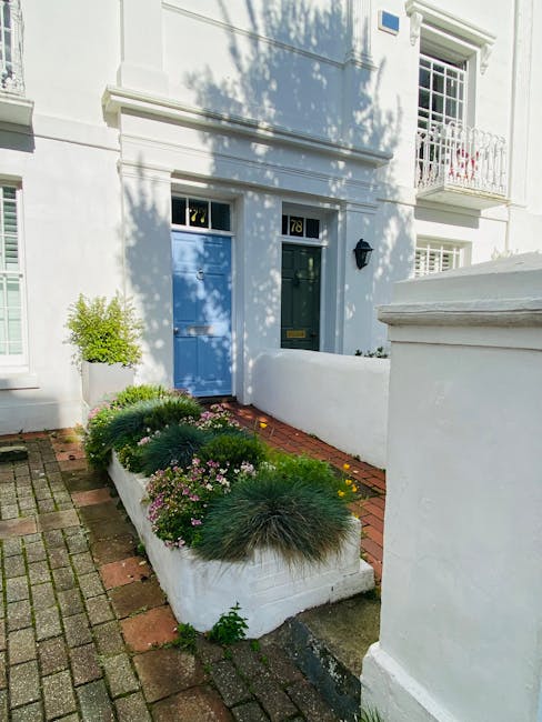 A small, neatly maintained front garden in a residential area with a white-painted boundary wall and a paved pathway leading to a light blue front door, displaying the house number 78. The garden feat