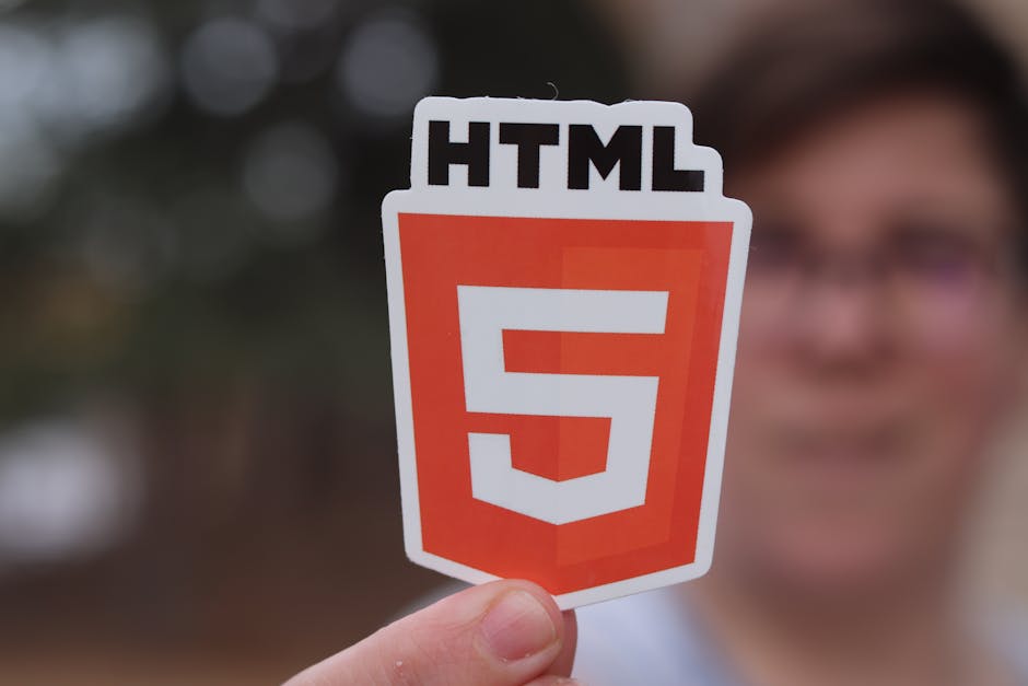 A person holding a small, colorful sticker with the HTML5 logo featuring a shield shape in orange and white, and the text 'HTML' at the top. The background is blurred with indistinct outdoor scenery a
